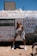 A person stands confidently in front of a weathered trailer with religious messages painted across it. The sky is clear and bright, and the individual is dressed in casual clothing, including sneakers, shorts, and a jacket draped off their shoulders.