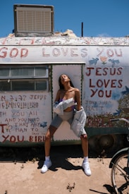 A person stands confidently in front of a weathered trailer with religious messages painted across it. The sky is clear and bright, and the individual is dressed in casual clothing, including sneakers, shorts, and a jacket draped off their shoulders.