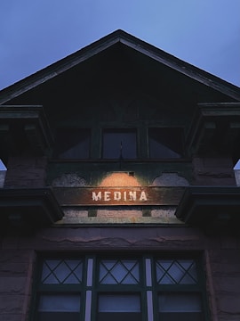 A building facade with a decorative triangular gable, featuring a light that illuminates the word 'MEDINA' centered on a wooden board. The structure has a rustic appearance with stone or brick elements and green-framed windows underneath. The sky above is overcast, suggesting a gloomy or late evening setting.