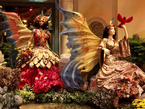 Two mannequin figures are styled as fantasy fairies, each adorned with large, colorful wings. The figure on the left wears a vibrant red and brown leaf dress embedded with flowers, while the figure on the right is dressed in a more subdued gold and brown gown, holding a red bird. Both are surrounded by lush greenery and natural elements, set in an elaborate display.