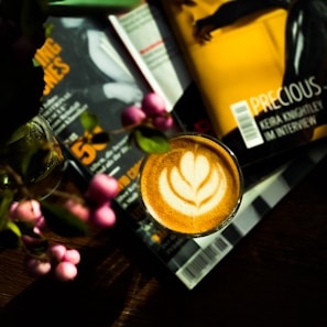 A cup of coffee with latte art featuring a leaf design sits on a table surrounded by books or magazines. Vibrant pink flowers and leaves partially obscure the view. The scene is well-lit, with natural light casting shadows and highlighting the textures of the coffee foam and the glossy surfaces of the reading material.