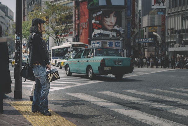 A busy urban intersection with a person standing on the sidewalk holding a shopping bag. A teal taxi is driving through the crosswalk, and a bus is visible in the background. Buildings with billboards and advertisements surround the area, and there are people walking on the opposite street.