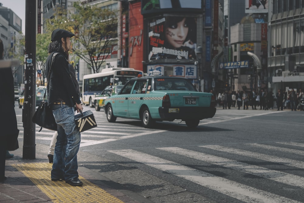 Mann in schwarzer Jacke und blauer Jeans steht tagsüber neben einem grünen Auto auf der Straße