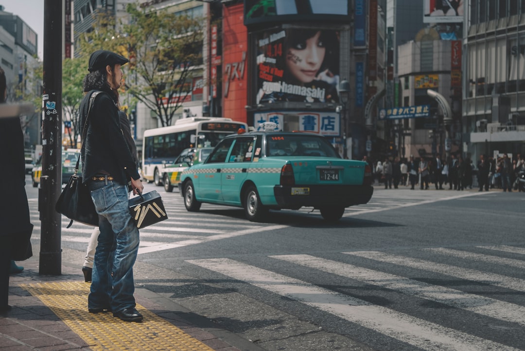 man in black jacket and blue denim jeans standing beside green car on road during daytime