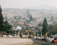 A small town nestled in a valley with a mixture of industrial and residential areas. An old mining structure stands prominently against a backdrop of hills, with rows of houses scattered throughout the landscape. A roadway lined with parked cars and a decorative archway sign can be seen in the foreground, surrounded by trees and greenery.