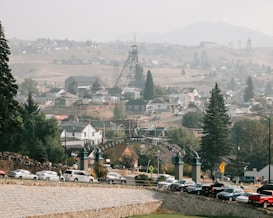 A small town nestled in a valley with a mixture of industrial and residential areas. An old mining structure stands prominently against a backdrop of hills, with rows of houses scattered throughout the landscape. A roadway lined with parked cars and a decorative archway sign can be seen in the foreground, surrounded by trees and greenery.