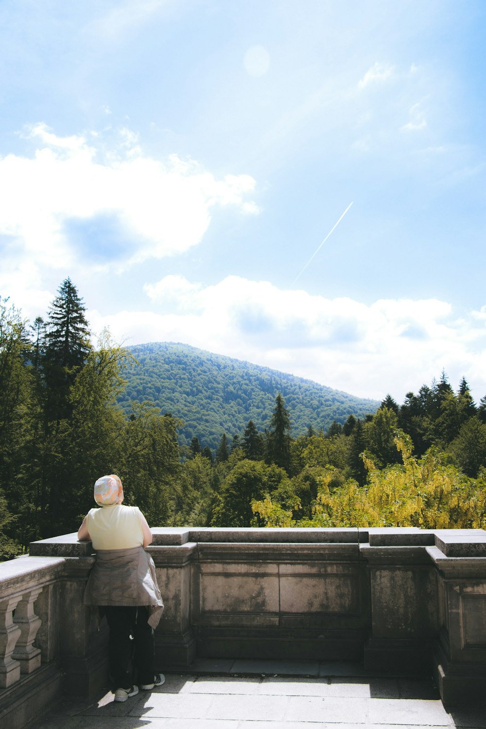 灰色の木製のベンチに座って昼間、緑の山々を眺める白いジャケットを着た女性