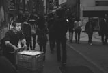 A man pushes a cart filled with items down a busy city street lined with shops and advertisements. Numerous pedestrians walk in various directions, creating a sense of bustling urban life.