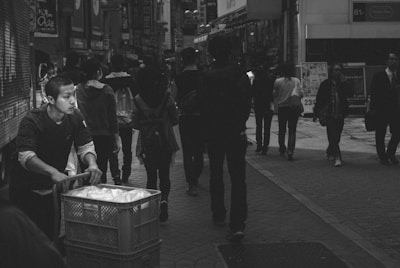 A man pushes a cart filled with items down a busy city street lined with shops and advertisements. Numerous pedestrians walk in various directions, creating a sense of bustling urban life.