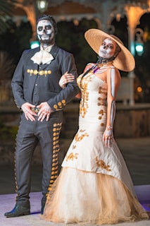 A couple is dressed in traditional Day of the Dead costumes with intricate face paint resembling skulls. The man is wearing a black charro suit with gold accents, while the woman is in a long, white dress adorned with gold patterns, wearing a wide-brimmed straw hat. They are posing together, arm in arm, in an outdoor setting with dim lights in the background.