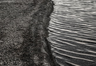 Dark sand transitions into gentle, rippling waves of water, creating a contrast between the rough texture of the sand and the smooth, wavy lines on the water surface.