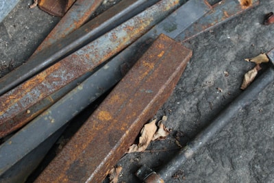 Rusted metal beams are scattered randomly on a rough concrete surface, with dry leaves strewn around.