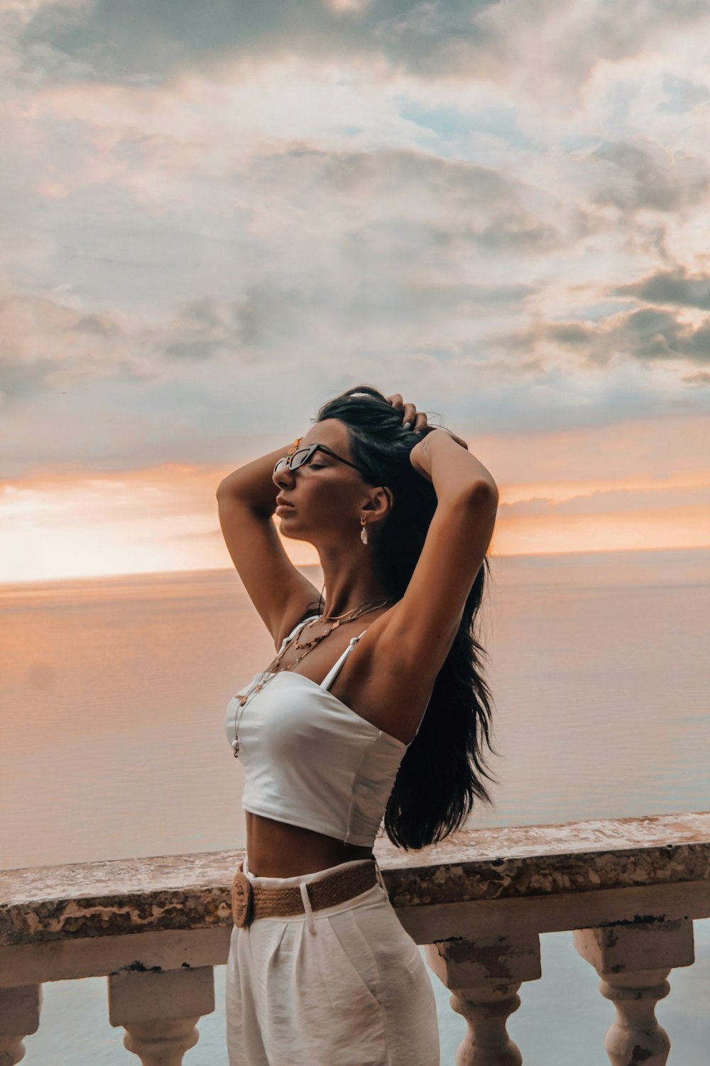 Mujer en camiseta blanca sin mangas y pantalones cortos de mezclilla azul de pie en la orilla de la playa durante la puesta del sol