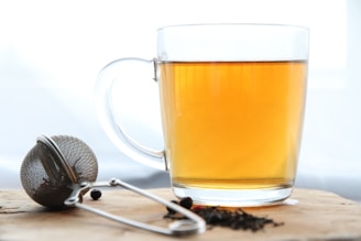 A clear glass cup filled with amber-colored tea is placed on a wooden surface. Beside it lies a metal tea infuser with some loose tea leaves scattered nearby.