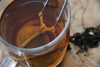 A close-up view of a glass cup filled with amber-colored tea. A metal tea infuser is submerged in the tea, attached to a handle with a clasp mechanism. Loose tea leaves are scattered on the wooden surface beside the cup.