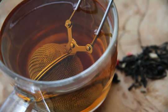 A close-up view of a glass cup filled with amber-colored tea. A metal tea infuser is submerged in the tea, attached to a handle with a clasp mechanism. Loose tea leaves are scattered on the wooden surface beside the cup.