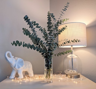 A decorative scene featuring a clear glass vase filled with tall eucalyptus branches placed on a light-colored tabletop. To the left, a white ceramic elephant figurine is displayed, and to the right, a lit table lamp with a beige shade provides soft lighting. Tiny string lights are scattered around the objects, adding a warm, cozy ambiance.