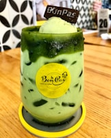 A green iced beverage served in a glass with ice cream and a chocolate piece on top. The glass is adorned with a yellow logo sticker and placed on a wooden table. The background shows a patterned wall and blurred cafe elements.
