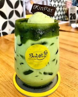 A green iced beverage served in a glass with ice cream and a chocolate piece on top. The glass is adorned with a yellow logo sticker and placed on a wooden table. The background shows a patterned wall and blurred cafe elements.