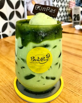 A green iced beverage served in a glass with ice cream and a chocolate piece on top. The glass is adorned with a yellow logo sticker and placed on a wooden table. The background shows a patterned wall and blurred cafe elements.