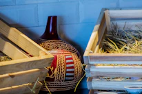 A large, ornately decorated ceramic vase with red and white patterns is nestled between two wooden crates filled with hay. The vase features intricate designs and motifs, adding a cultural touch to the rustic setting.