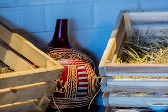 A large, ornately decorated ceramic vase with red and white patterns is nestled between two wooden crates filled with hay. The vase features intricate designs and motifs, adding a cultural touch to the rustic setting.