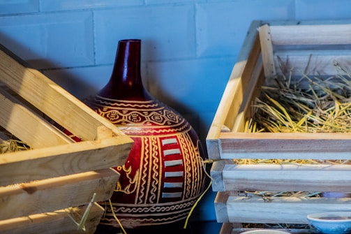 A large, ornately decorated ceramic vase with red and white patterns is nestled between two wooden crates filled with hay. The vase features intricate designs and motifs, adding a cultural touch to the rustic setting.