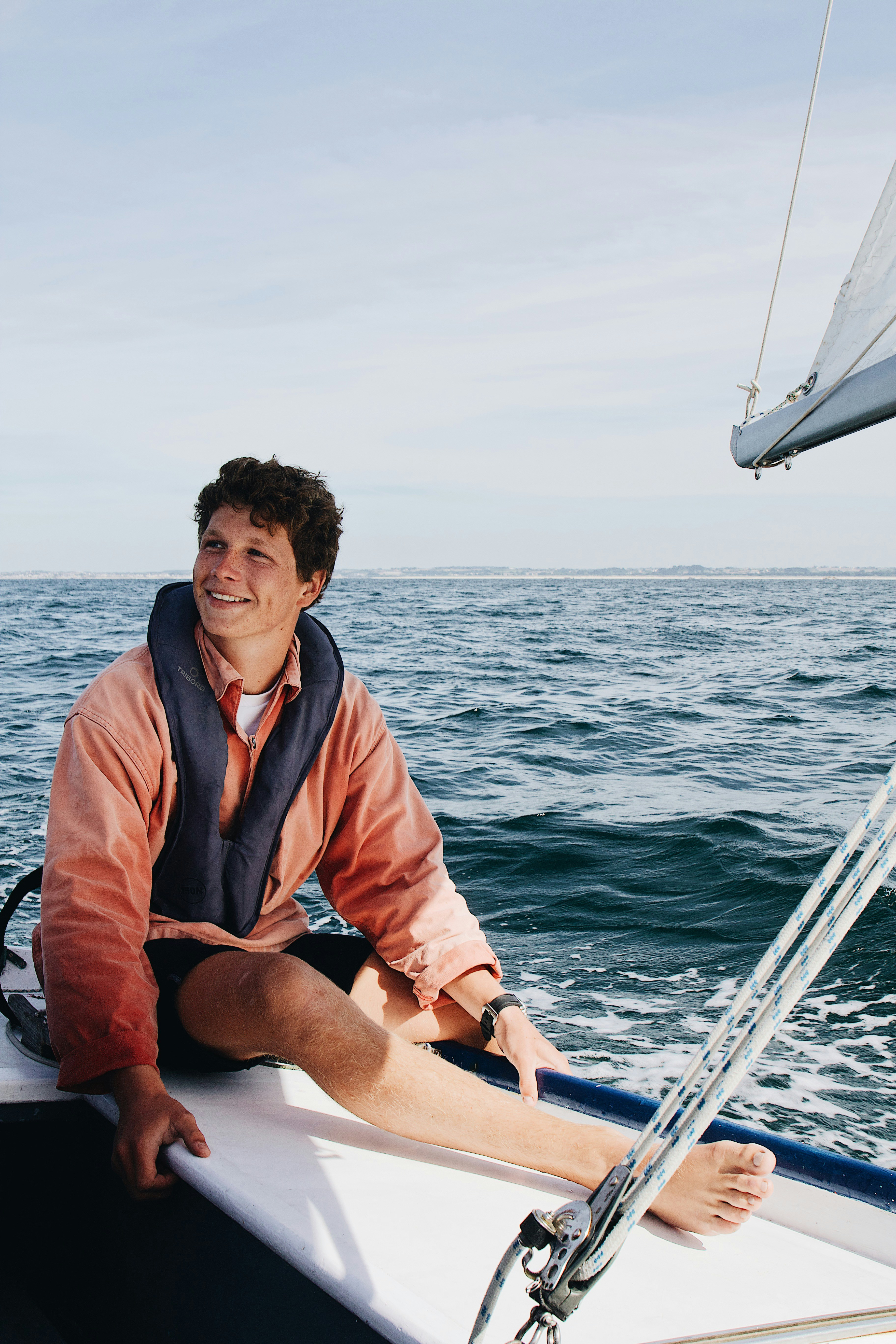Smiling young man in a salmon-colored jacket sitting on the edge of a sailboat at sea, a navy inflatable life jacket worn flat around his neck and chest