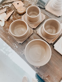 Several clay pottery pieces in progress are arranged on a wooden table. Various pottery tools and a sponge are scattered nearby, suggesting active work. The clay items appear to be in the form of bowls or cups, placed on square wooden bases.