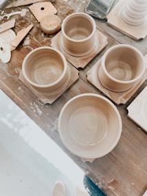 Several clay pottery pieces in progress are arranged on a wooden table. Various pottery tools and a sponge are scattered nearby, suggesting active work. The clay items appear to be in the form of bowls or cups, placed on square wooden bases.