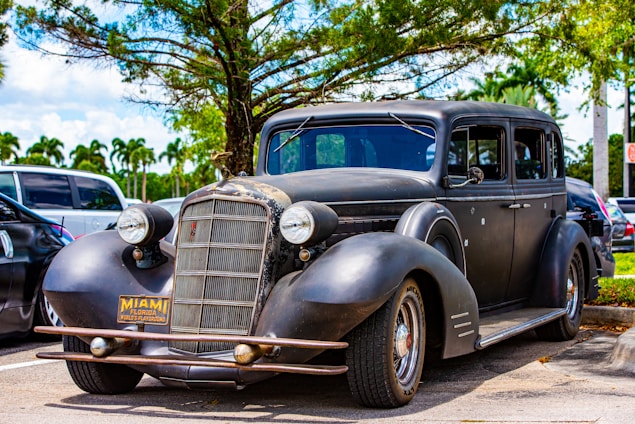 A vintage black car with a rusted front grill parked under a tree. The car bears a Miami, Florida license plate and is situated in a parking lot with other vehicles. The surrounding area features palm trees and a clear blue sky, indicating a sunny day.