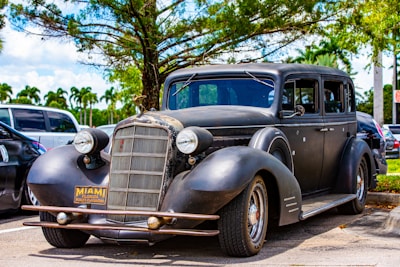 A vintage black car with a rusted front grill parked under a tree. The car bears a Miami, Florida license plate and is situated in a parking lot with other vehicles. The surrounding area features palm trees and a clear blue sky, indicating a sunny day.