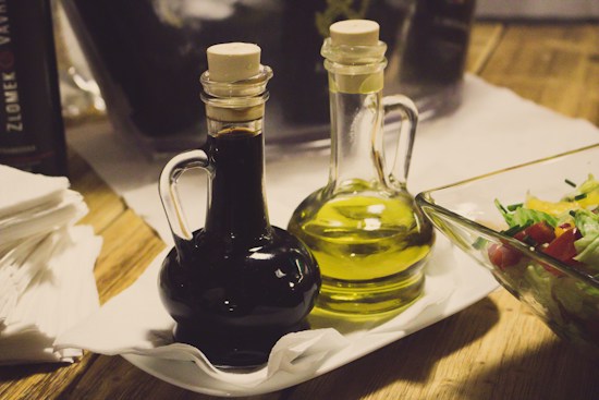 A wooden table setting featuring a white rectangular plate holding two glass bottles with cork stoppers, one containing a dark liquid and the other a light yellow liquid. Next to the bottles is a glass bowl filled with a fresh salad. Napkins can be seen stacked on the side.