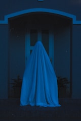 A person draped in a white sheet stands in front of a doorway, creating a ghostly figure against a dimly-lit backdrop. The scene conveys a mysterious and slightly eerie atmosphere with shadows adding to the intrigue.