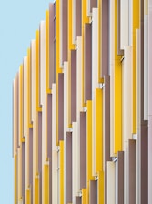 Vertical panels in alternating shades of yellow, beige, and brown create a modern, geometric facade against a pale blue background. The repetitive pattern draws the eye upward, showcasing a contemporary architectural design.