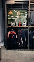 A store display features a large screen showcasing a man in a grey hoodie with the Adidas logo, while below it are two Adidas soccer balls and a red bottle. The clothing rack beneath holds hanging Adidas jackets, including a prominent navy blue and red jacket with white stripes, alongside dark-colored track pants.