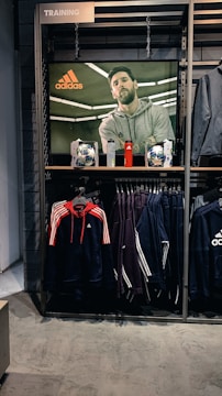 A store display features a large screen showcasing a man in a grey hoodie with the Adidas logo, while below it are two Adidas soccer balls and a red bottle. The clothing rack beneath holds hanging Adidas jackets, including a prominent navy blue and red jacket with white stripes, alongside dark-colored track pants.