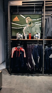A store display features a large screen showcasing a man in a grey hoodie with the Adidas logo, while below it are two Adidas soccer balls and a red bottle. The clothing rack beneath holds hanging Adidas jackets, including a prominent navy blue and red jacket with white stripes, alongside dark-colored track pants.