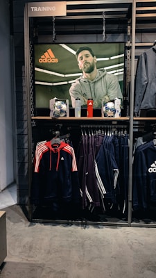 A store display features a large screen showcasing a man in a grey hoodie with the Adidas logo, while below it are two Adidas soccer balls and a red bottle. The clothing rack beneath holds hanging Adidas jackets, including a prominent navy blue and red jacket with white stripes, alongside dark-colored track pants.