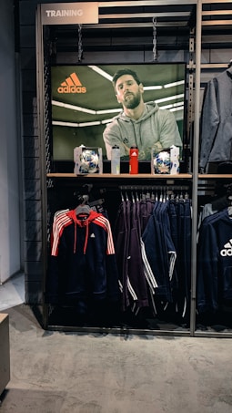 A store display features a large screen showcasing a man in a grey hoodie with the Adidas logo, while below it are two Adidas soccer balls and a red bottle. The clothing rack beneath holds hanging Adidas jackets, including a prominent navy blue and red jacket with white stripes, alongside dark-colored track pants.