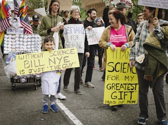 A group of people participating in a march or protest hold various signs advocating for science. One child, dressed in a white lab coat, carries a sign that reads 'Without Science Bill Nye is Just The Guy.' Another person holds a sign stating 'Thank Science for My Greatest Gift Proud IVF Mom.' Several rainbow and American flags are displayed on a cart filled with buttons. The crowd appears engaged and supportive of scientific causes.