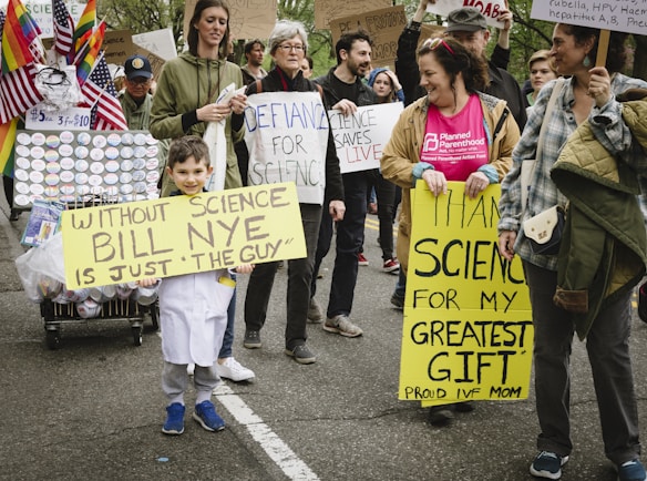 A group of people participating in a march or protest hold various signs advocating for science. One child, dressed in a white lab coat, carries a sign that reads 'Without Science Bill Nye is Just The Guy.' Another person holds a sign stating 'Thank Science for My Greatest Gift Proud IVF Mom.' Several rainbow and American flags are displayed on a cart filled with buttons. The crowd appears engaged and supportive of scientific causes.