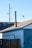 A wooden fence stands in front of an old building with a sloped roof. A tall metal chimney rises from the roof. Visible are electrical wires and a light pole in the background against a clear blue sky.