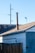 A wooden fence stands in front of an old building with a sloped roof. A tall metal chimney rises from the roof. Visible are electrical wires and a light pole in the background against a clear blue sky.