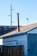 A wooden fence stands in front of an old building with a sloped roof. A tall metal chimney rises from the roof. Visible are electrical wires and a light pole in the background against a clear blue sky.