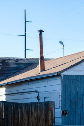 A wooden fence stands in front of an old building with a sloped roof. A tall metal chimney rises from the roof. Visible are electrical wires and a light pole in the background against a clear blue sky.