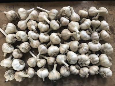 A collection of garlic bulbs is spread out on a wooden surface. The bulbs are arranged closely together, showcasing their papery white skins and dried brown roots.