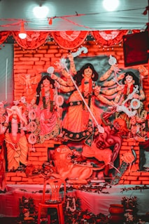 A vibrant display featuring a sculptural depiction of Hindu deities adorned with intricate costumes and ornaments. The figures are positioned against a brick-like backdrop, with bright red and orange tones dominating the scene. Decorative elements such as flowers, traditional motifs, and garlands surround the deities. The central figure holds multiple weapons, symbolizing power and divine energy.