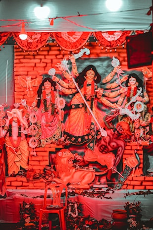 A vibrant display featuring a sculptural depiction of Hindu deities adorned with intricate costumes and ornaments. The figures are positioned against a brick-like backdrop, with bright red and orange tones dominating the scene. Decorative elements such as flowers, traditional motifs, and garlands surround the deities. The central figure holds multiple weapons, symbolizing power and divine energy.