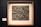 A framed artwork featuring a textured, abstract sculpture with various rectangular and geometric shapes. The piece has a three-dimensional quality with rough, uneven surfaces and a metallic appearance.